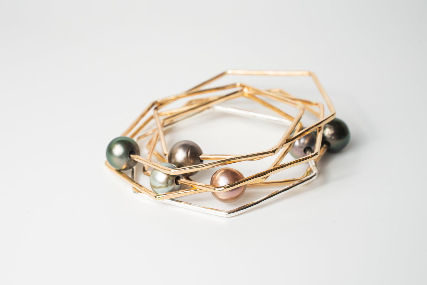 Stacked Lōkahi gold-filled geometric bangles featuring mixed-tone Tahitian pearls in green, silver, and blush hues on a white background.