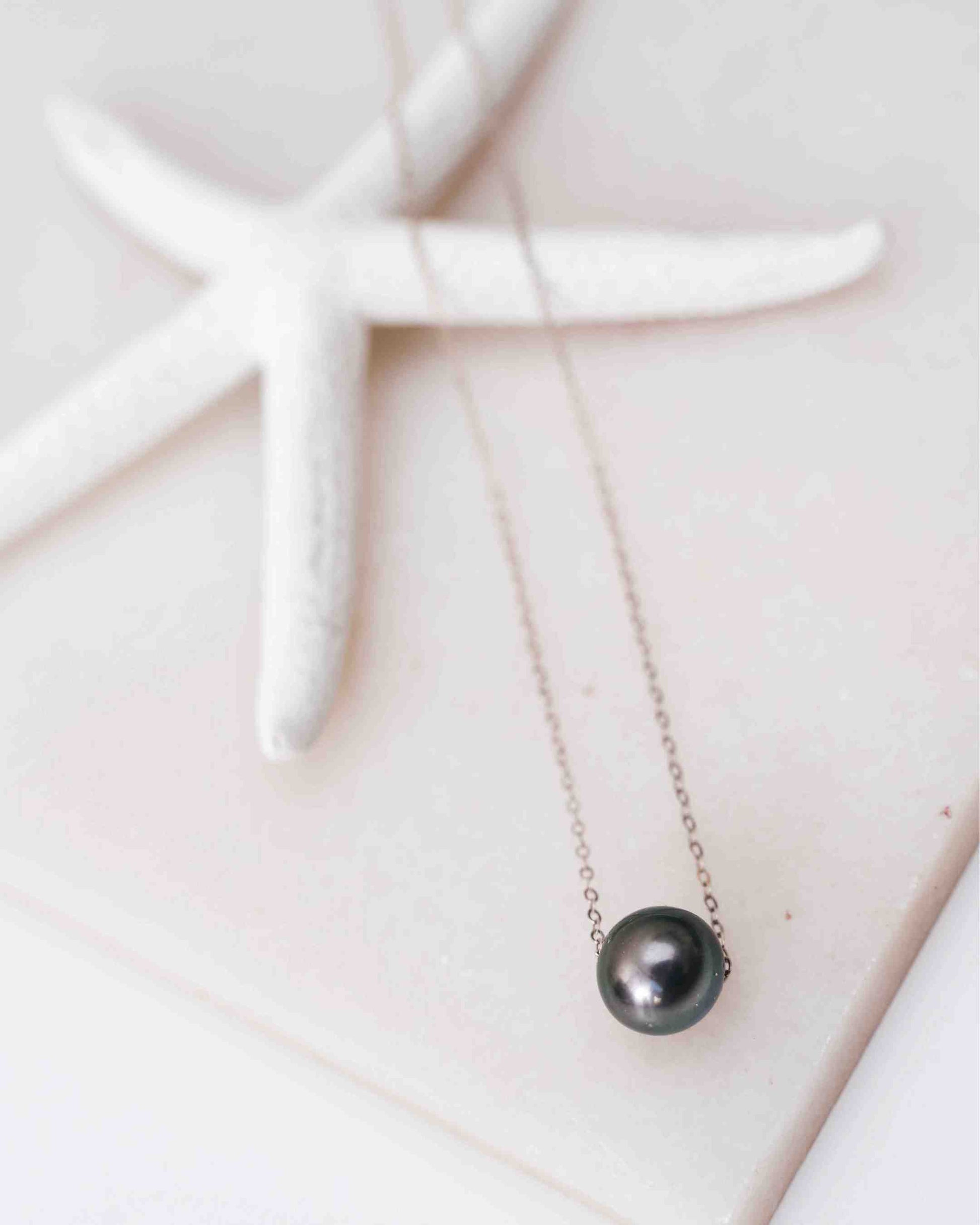 The Mo‘i Necklace with a single Black Tahitian pearl on a fine silver chain, styled on a white starfish background.
